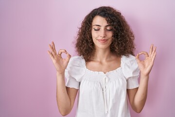 Hispanic woman with curly hair standing over pink background relax and smiling with eyes closed doing meditation gesture with fingers. yoga concept.