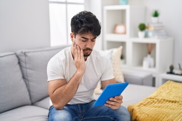 Hispanic man with beard using touchpad sitting on the sofa touching mouth with hand with painful expression because of toothache or dental illness on teeth. dentist concept.