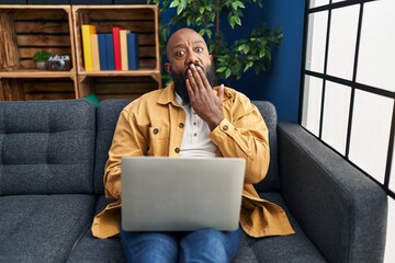African american man using laptop at home sitting on the sofa covering mouth with hand, shocked and afraid for mistake. surprised expression