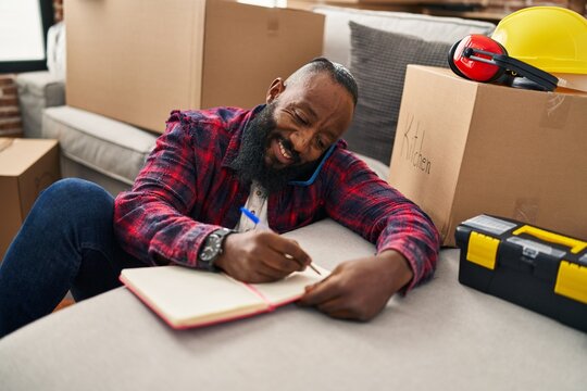 Young african american man talking on the smartphone and writing on book sitting on floor at new home