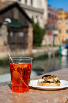 Aperol spritz cocktail in plastic cup with cicchetti snack by canal in Venice Italy sunny travel street food scene