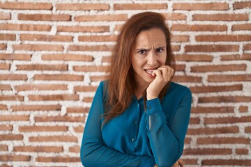 Brunette woman standing over bricks wall looking stressed and nervous with hands on mouth biting nails. anxiety problem.