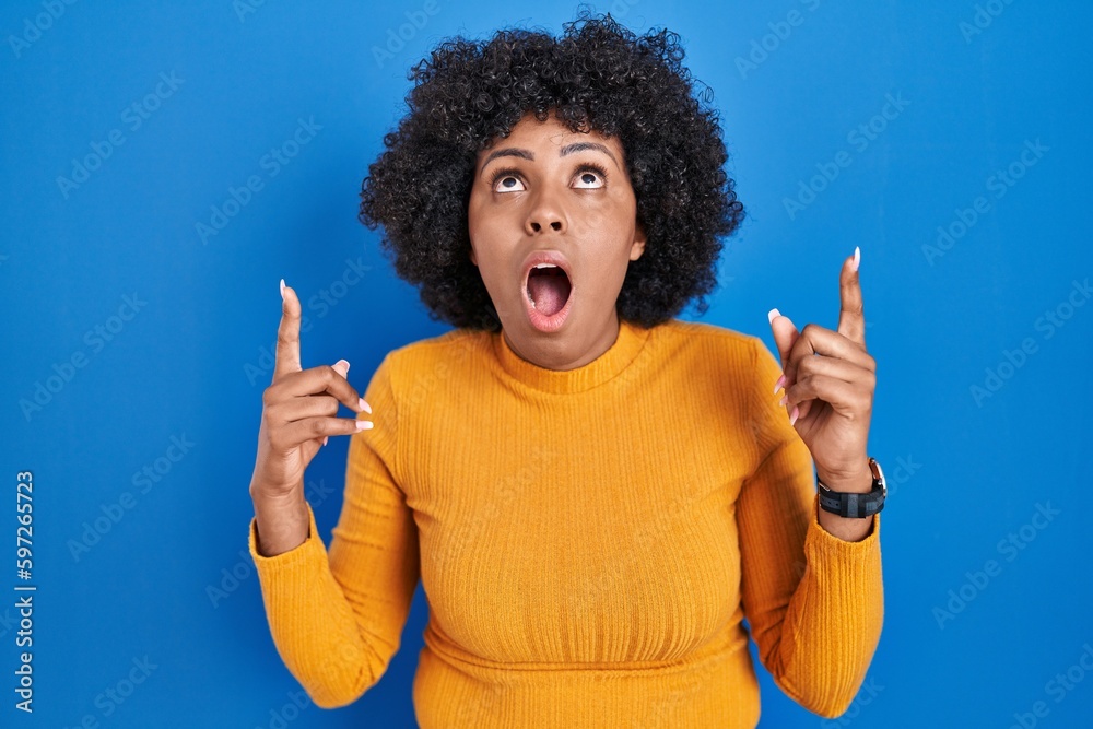 Poster black woman with curly hair standing over blue background amazed and surprised looking up and pointi