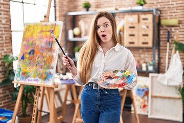 Young caucasian woman at art studio holding palette scared and amazed with open mouth for surprise,...
