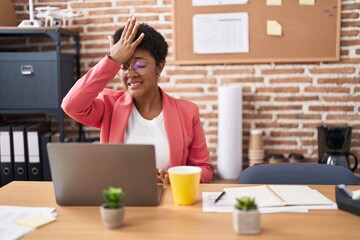 Young african american woman working at the office wearing glasses surprised with hand on head for...