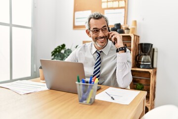 Middle age grey-haired man business worker using laptop talking on smartphone at office