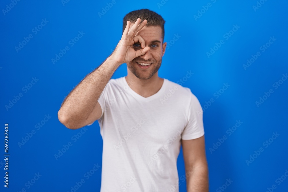 Poster Young caucasian man standing over blue background doing ok gesture with hand smiling, eye looking through fingers with happy face.