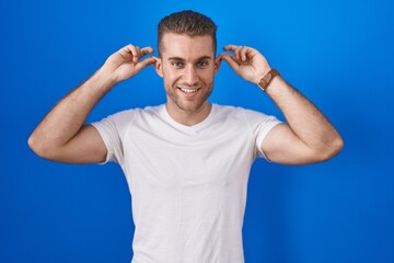 Young caucasian man standing over blue background smiling pulling ears with fingers, funny gesture. audition problem