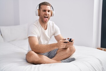 Young caucasian man sitting on the bed at home playing video games sticking tongue out happy with...