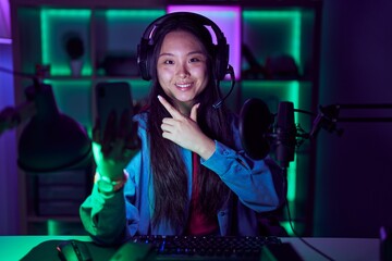 Young asian woman playing video games with smartphone cheerful with a smile on face pointing with hand and finger up to the side with happy and natural expression