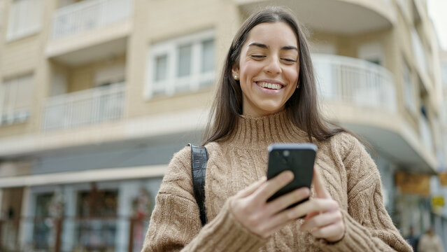 Young beautiful hispanic woman using smartphone smiling at street