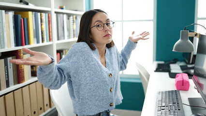 Young beautiful hispanic woman student sitting on table with i dont know expression at library...