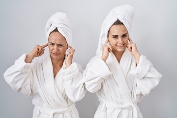 Middle age woman and daughter wearing white bathrobe and towel covering ears with fingers with annoyed expression for the noise of loud music. deaf concept.