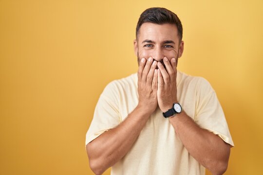 Handsome hispanic man standing over yellow background laughing and embarrassed giggle covering mouth with hands, gossip and scandal concept