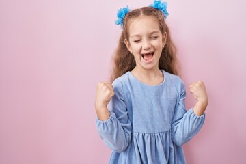 Young little girl standing over pink background very happy and excited doing winner gesture with arms raised, smiling and screaming for success. celebration concept.