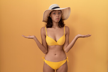 Young hispanic woman wearing bikini and summer hat clueless and confused expression with arms and hands raised. doubt concept.