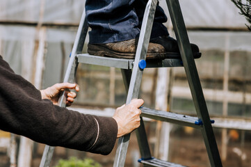 A woman assistant holds an aluminum stepladder with her hands, insuring a worker, a man, a builder in a dirty robe standing on the steps. Photography, portrait of legs, shoe close-up, lifestyle. © shchus