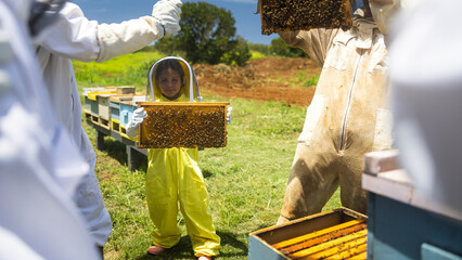 Beekeeper kid in a protective suit holding a honeycomb with bees from the beehive at the apiary.