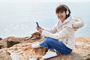 Middle age woman listening to music sitting on the rock at seaside
