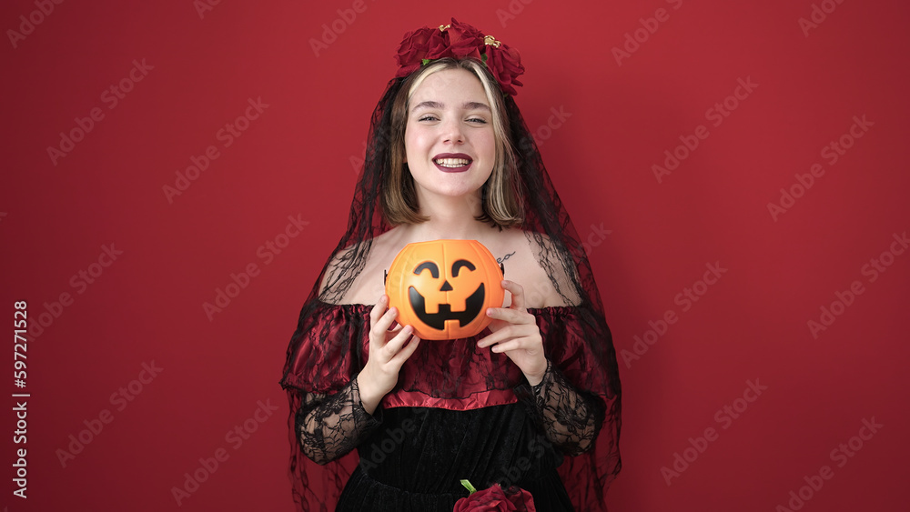 Wall mural Young blonde woman wearing katrina costume holding halloween pumpkin basket over isolated red background