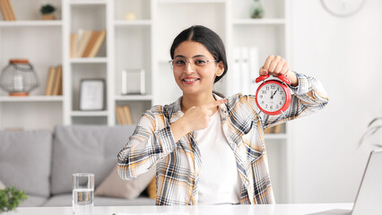 Indian smiling woman showing time on clock, approving, pointing finger at clock, encouraging. This...