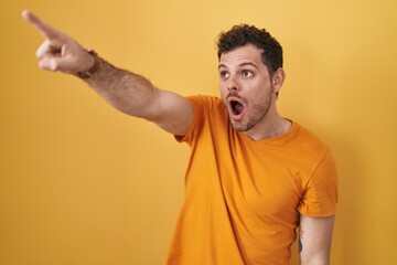 Young hispanic man standing over yellow background pointing with finger surprised ahead, open mouth amazed expression, something on the front