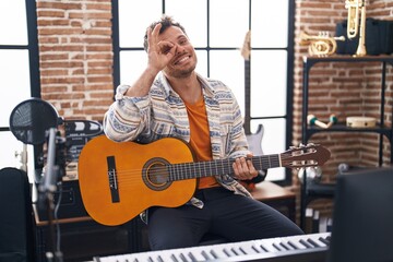 Young hispanic man playing classic guitar at music studio smiling happy doing ok sign with hand on eye looking through fingers