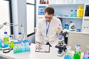 Young hispanic man scientist holding test tube write on document at laboratory