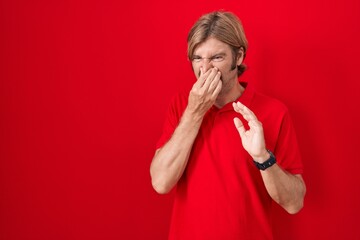 Caucasian man with mustache standing over red background smelling something stinky and disgusting, intolerable smell, holding breath with fingers on nose. bad smell