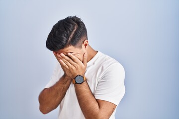 Hispanic man standing over blue background with sad expression covering face with hands while...