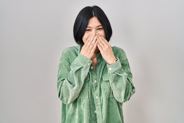 Young asian woman standing over white background laughing and embarrassed giggle covering mouth...