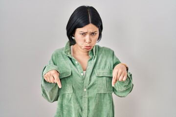 Young asian woman standing over white background pointing down looking sad and upset, indicating...