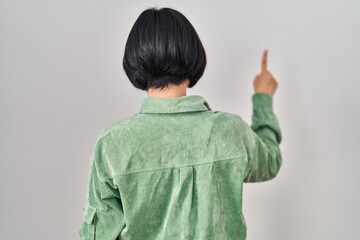 Young asian woman standing over white background posing backwards pointing ahead with finger hand