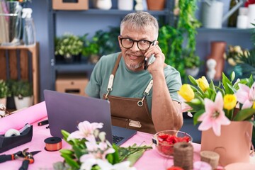 Middle age grey-haired man florist talking on smartphone using laptop at flower shop