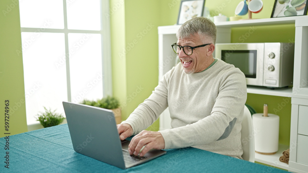 Poster middle age grey-haired man using laptop sitting on table at home