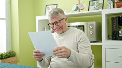Middle age grey-haired man reading document sitting on table at home