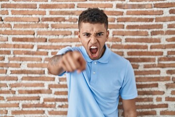 Brazilian young man standing over brick wall pointing displeased and frustrated to the camera, angry and furious with you