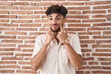 Arab man with beard standing over bricks wall background smiling with open mouth, fingers pointing and forcing cheerful smile