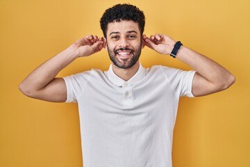 Arab man standing over yellow background smiling pulling ears with fingers, funny gesture. audition...
