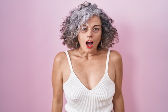 Middle age woman with grey hair standing over pink background afraid and shocked with surprise and amazed expression, fear and excited face.