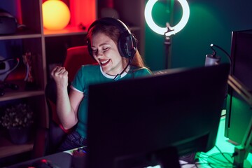 Redhead woman playing video games very happy and excited doing winner gesture with arms raised, smiling and screaming for success. celebration concept.