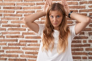 Young caucasian woman standing over bricks wall doing bunny ears gesture with hands palms looking cynical and skeptical. easter rabbit concept.