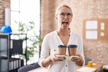 Young caucasian woman working at the office holding coffee cups angry and mad screaming frustrated and furious, shouting with anger looking up.