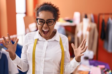 Young hispanic woman with curly hair standing at tailor room crazy and mad shouting and yelling...