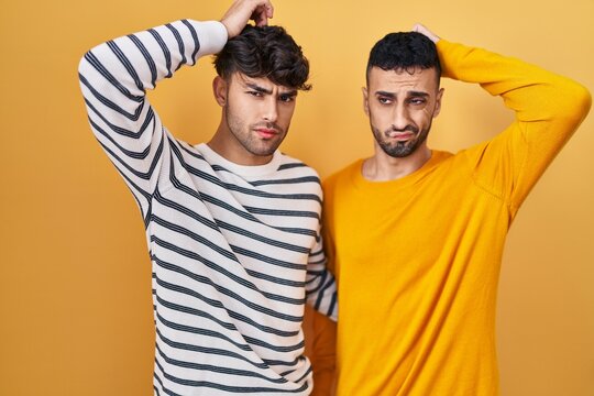 Young hispanic gay couple standing over yellow background confuse and wondering about question. uncertain with doubt, thinking with hand on head. pensive concept.