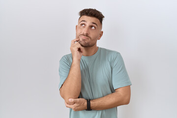 Hispanic man with beard standing over white background with hand on chin thinking about question, pensive expression. smiling with thoughtful face. doubt concept.