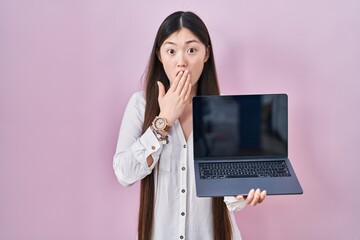 Chinese young woman holding laptop showing screen covering mouth with hand, shocked and afraid for...