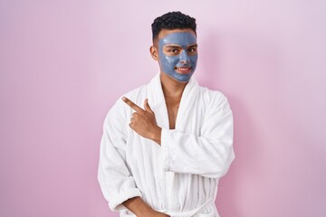 Young hispanic man wearing beauty face mask and bath robe cheerful with a smile on face pointing with hand and finger up to the side with happy and natural expression