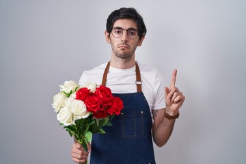 Young hispanic man holding bouquet of white and red roses pointing up looking sad and upset,...
