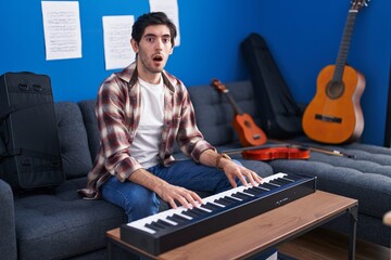 Young hispanic man playing piano at music studio in shock face, looking skeptical and sarcastic, surprised with open mouth © Krakenimages.com